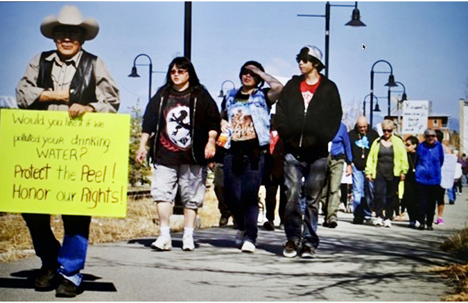 A collection of protesters, young and old, Indigenous and non-Indigenous people walk along a sidewalk during a day-time march. Some are carrying signs that share the messages: PROTECT THE PEEL, HONOUR OUR RIGHTS and other statements.