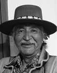 Black and white photograph: Portrait of a man with a moustache smiling and wearing a hat with decorated band, a patterned collared shirt, and a collared coat. 