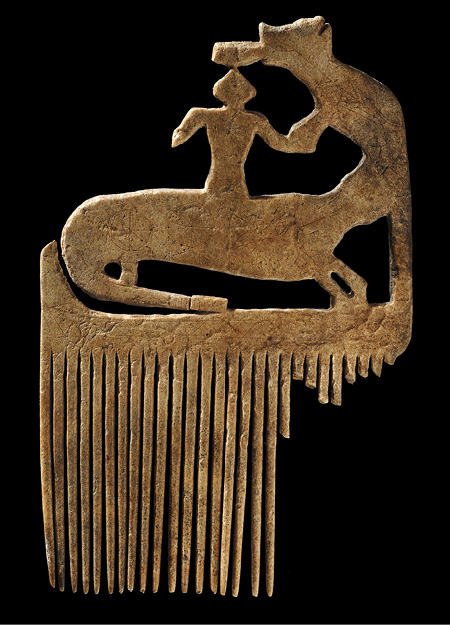 Colour Photograph. Photo of a seventeenth century moose antler comb. The comb is a golden brown colour, and has a carving of a human-like figure and two animals at the top. The teeth of the comb descend from this, although the rightmost teeth are all broken.