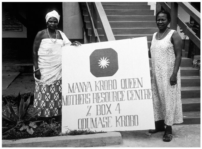 A black and white photograph of two women from Manya Krobo Queenmothers Association, holding a banner with text Manya Krobo Queenmothers Resource Centre, Box 4, Odumase Krobo outside the Resource Centre office.