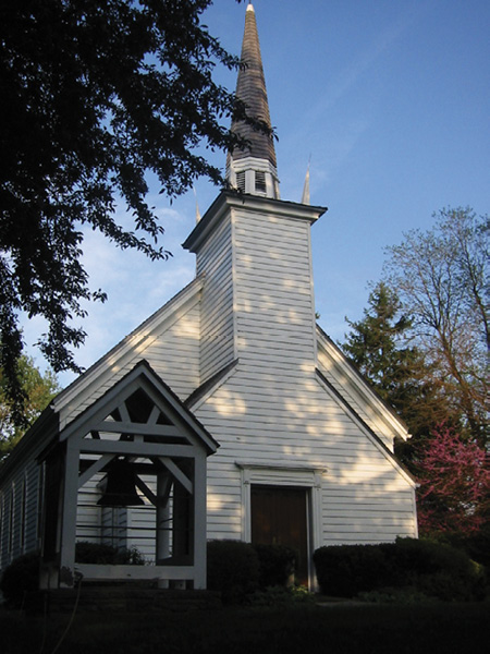 Colour photograph. A white clapboard church with a tall steeple and a small front door. There is a roofed wooden frame housing a large cast iron bell in front of the church on the left.