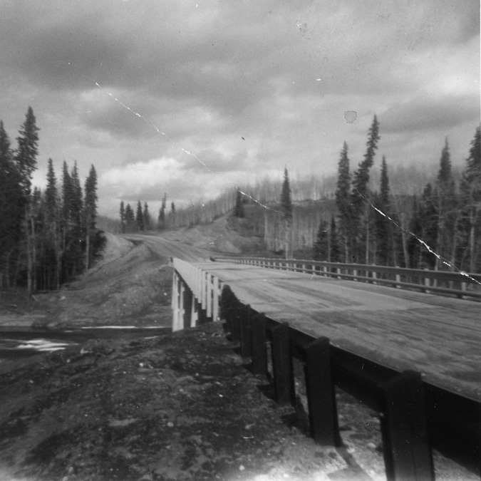The image is a black and white photograph depicting a rural landscape with a narrow bridge crossing over a river. The bridge extends diagonally from the bottom right towards the center-left, with wooden railings on both sides. Tall coniferous trees densely line the dirt road on the left side of the bridge, stretching into the distance. A dirt road, visibly rough and uneven, approaches the bridge from the left. In the background are more trees silhouetted against a cloudy sky, adding depth to the scene.