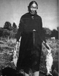 Black and white photograph: Older photograph of a woman standing outside on a sunny day in a calve-length dress holding a fish in each hand.
