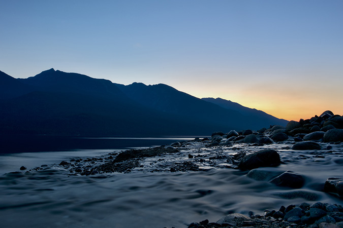 A still body of water in the middle of the view is bookended with many small and large shoreline rocks resting in the foreground. A rounded mountain sits in the background with a dark and cloudy sky above. The mood is quiet but foreboding. This image was gathered where the Silverton Creek empties into Lake Slocan in British Columbia.