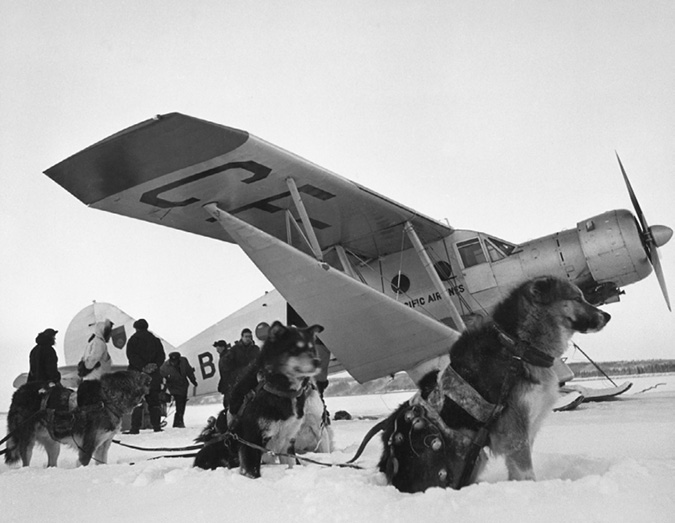 This photo shows a team of husky sled dogs sitting in front of a single engine seaplane with several men in the background. Because of weather conditions in the winter and spring, dog teams, canoes and planes were the main modes of transportation in the Northwest Territory in this era.