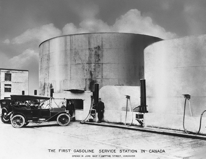 This photo shows two automobiles parked next to oil storage tanks. A man stands beside a gas pump in front of the storage tank next to one of the automobiles. This was later heralded as the first “service station” by Imperial Oil.