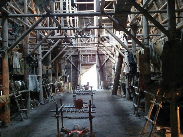 Colour photograph. Photo of the interior of a reconstructed longhouse. Complex wall and ceiling beams run along the length of the longhouse, with an open door visible at the back. Two firepits, with small wooden racks above them, are in the centre of the building. A variety of ladders are placed on the sides of the walls, leading to sleeping platforms, and skins and other materials are hanging off the platforms and beams.