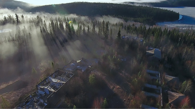 An aerial view of Uranium City buildings and structures, now abandoned, and blending into the landscape of evergreen trees in the foreground and mountains touched with low hanging cloud and mist.
