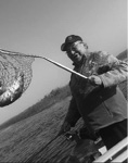 Black and white photograph: Photograph of a man in a boat on a lake, wearing a ball cap, holding a freshly caught fish in a net. 