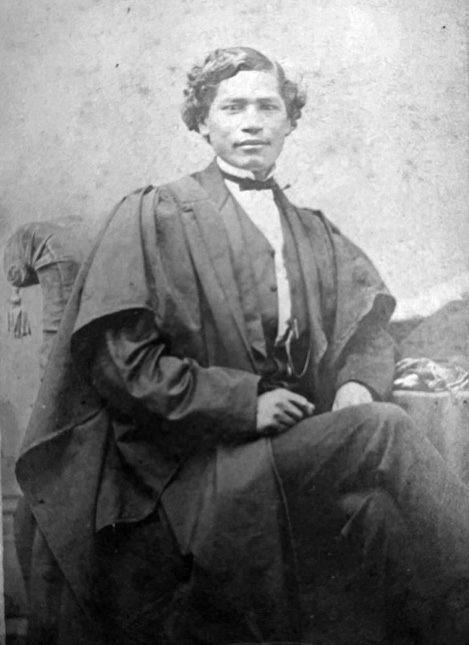 Black and white photograph: Indigenous man in his twenties or thirties with short curly black hair sitting indoors in a formal pose on a chair. This appears to be a photograph taken by a professional photographer.  He is formally dressed with a bow tie and a shirt with an upright collar and a robe.