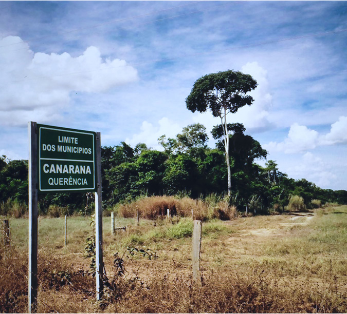 . A view of a fence and signage at the border of two municipalities of the Upper Xingu Basin (Canarana and Querência).  The grassland, arid Cerrado and lush forest biomes are on either side of the sign and fence. The grassland to the right of the sign and in the image, the forest behind the sign to the left of the frame.