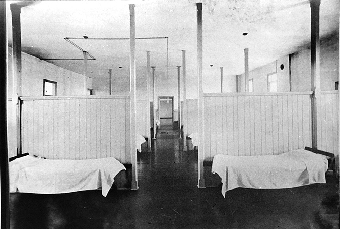 Black and white photograph: Dormitory showing a very simple unadorned room with rows of single beds separated by dividers. It looks very clinical, almost like a hospital.