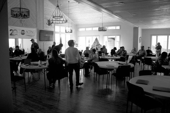 Black and white photograph: A large, sunny room where a group of adults are gathered and socializing. Most individuals are sitting at various round tables, and a few are standing. Dark, modern chandeliers hang from the wood-paneled ceiling.  A small white tipi with designs painted on it stands in front of the large window at the back of the room. A large banner hangs from the far-left wall, which reads 20 years ACFN.