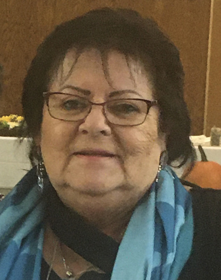 Colour photograph. Head and shoulders portrait of an Indigenous woman with mid-length dark hair and wearing glasses. She is also wearing pendant earrings and a blue scarf.
