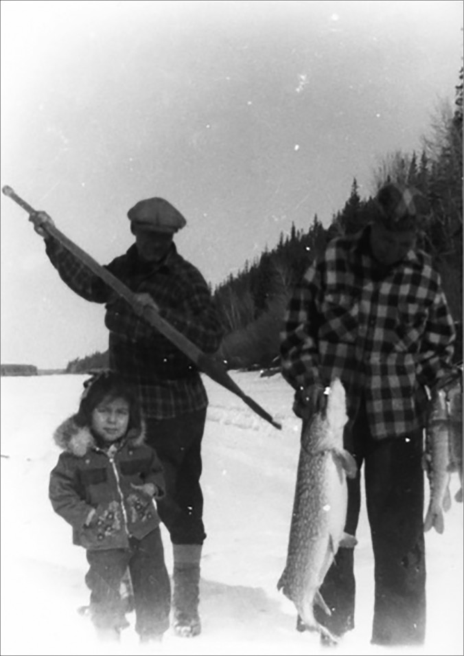 The black and white photograph depicts three people standing in a snowy outdoor setting, possibly a frozen river or lake. In the foreground, a child (Mary Beaver) wearing a winter coat with a fur-lined hood stands facing the camera, appearing to smile. Behind the child, two older individuals (Felix and Mary Ann Beaver), both in checkered jackets and winter hats, hold fishing spears and large fish. The person on the left holds a spear upright over their shoulder, while the person on the right holds two large fish. In the background, a line of trees is visible, silhouetted against the sky.