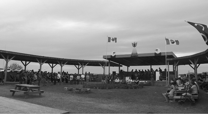 Black and white photograph: An outdoor gathering of children, middle-aged and elderly people. They are gathered near a large, round canopy structure supported by wooden posts and open to the sky in the centre. It is an overcast day. In order from left to right, the Canadian flag, ACFN’s flag and the Métis infinity symbol flag are blowing in the wind.  In the foreground is an empty picnic table and an unlit stone fire pit. To the left are people standing in a line. In the center Elders are sitting in chairs. To the right are people sitting at picnic tables. In the background, behind the seated Elders, are a line of male drummers standing on a stage behind microphones. Above the stage, the canopy roof is decorated with two large painted eagle heads and a large, painted grey and white canvas lodge.