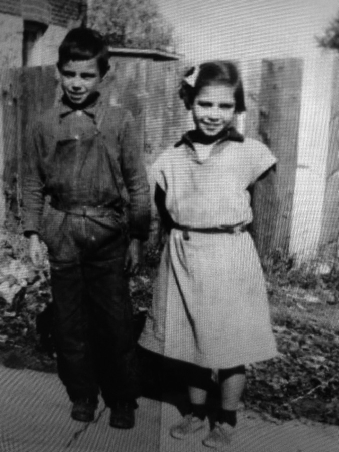 Black and white photograph: Young Indigenous boy around eleven years of age dressed in overalls, standing beside his sister, who is two years younger.  The boy is dressed in overalls and the girl in a loose-fitting dress.  They are outside and both are smiling.