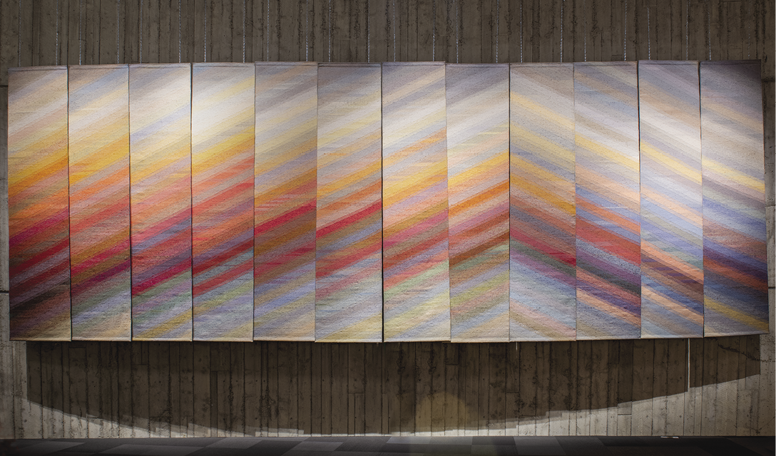 Photo: a series of twelve tall hangings that from a pyramid pattern from orange, red, pin, and yellow lines against a blue-grey background. 