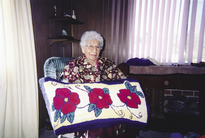 Grey haired woman in a wood-panelled room holds up a carpet with three red poppies on it.