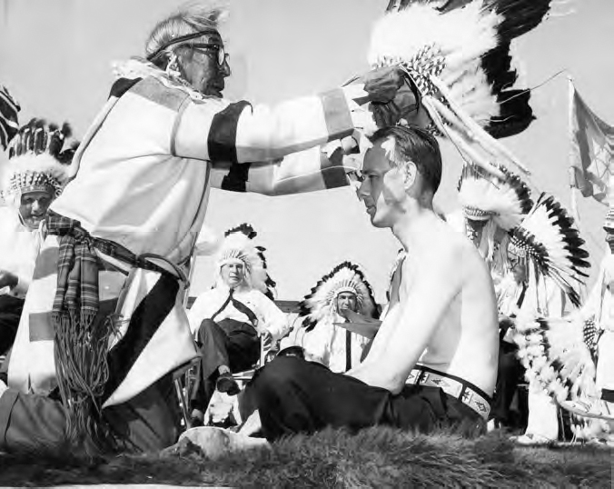 A photograph shows an old man wearing a traditional dress, putting a crown on a young man's head. Several other people wearing the same traditional dress can be seen in the background.