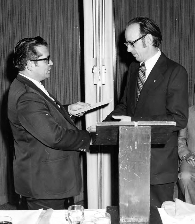 A photograph shows two men wearing formal dresses with spectacles, exchanging some notes. A dark shaded wall can be seen in the background.