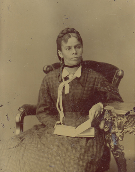 Black and white historical photograph. Portrait of an Indigenous woman seated on a large upholstered chair with carved wooden armrests. She is wearing a checked dress and a white neck tie, and is holding an open book on her lap while looking to the right.