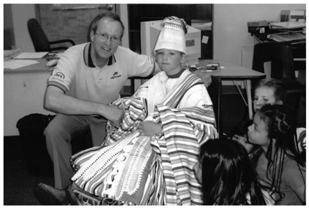 A black and white photograph of Brian Keating, a young man with Brett Prindle, a toddler dressed in African traditional clothing. Other young children are sitting around them.