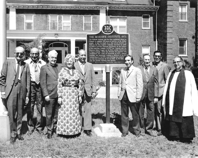 Black and white photograph: Group of male and female adults standing on both sides of a metal plaque on a stand in front of the Mohawk Institute.  On the left side of the photograph are three men in suits, one man dressed as a priest, and a woman in a long dress.  On the right side of the photograph there are three men in suits and a man in religious garb/robe. The text of the plaque is unreadable but the plaque title reads “THE MOHAWK INSTITUTE 1831.”  The coat of arms for Ontario is at the top of the plaque.