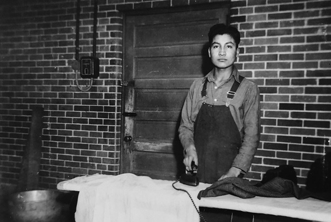 Black and white photograph: Teenage Indigenous boy in overalls and a long-sleeve work shirt ironing in front of a brick wall.