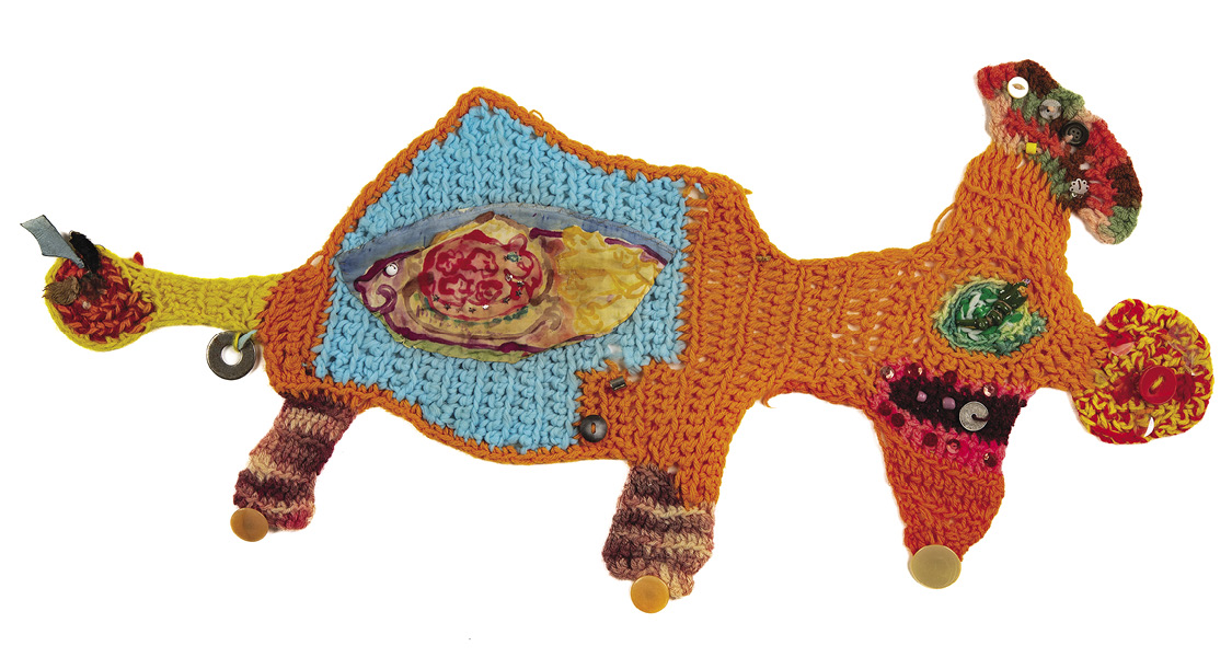 Photo: primarily orange, strangely shaped knitted “creature” with four legs, a yellow tail, buttons, splashes of brown, red, blue, and a section in the middle made from a different materials and looking almost like a fish.