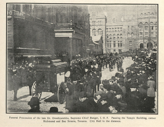 Black and white historical photograph. A funeral procession on a downtown street lined on both sides by a large crowd of onlookers. The horse-drawn funeral carriage is closest to the viewer, with an honour guard and other carriages or early cars leading the procession. Elaborate brick buildings are visible at the end of the street, and a typed caption is visible underneath the photograph.