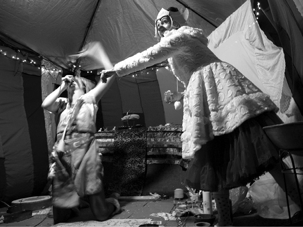 Black and white photograph, two people perform inside an eclectically decorated tend, the performer on the left is singing and dancing with their movements blurred by the photograph, the performer on the right dances while wearing a thick polka dot dress, a clown nose, and a cardboard crown.