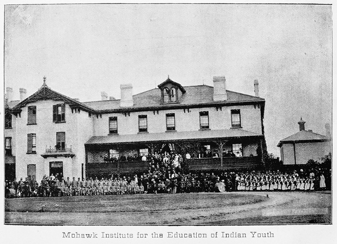 Black and white photograph of postcard: Second set of Mohawk Institute buildings.  Pictured is a two-storey brick building (and lower level) with a large porch across the front and an addition on the left side of the main building.  Students, teachers and others cover the lawn in front of the school and the school steps.  There are over 200 people in the photograph. The caption on the image reads “Mohawk Institute for the Education of Indian Youth.”