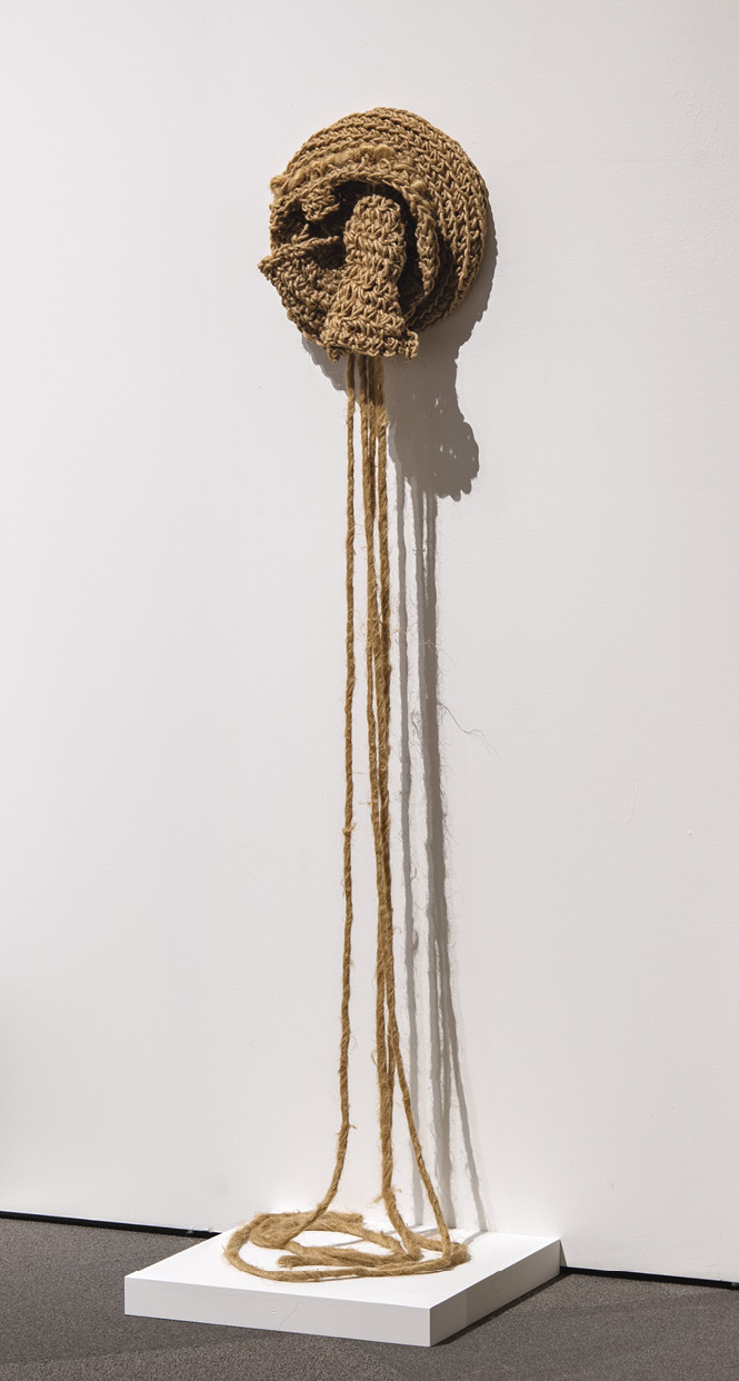 Photo: a circle of light brown knitted fabric sits on a white wall with an opening disgorging ropes to a white shelf on the grey cement floor.