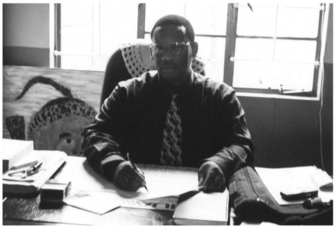 A black and white photograph of inkosi Bhengu, sitting at his desk in his office, doing paperwork.