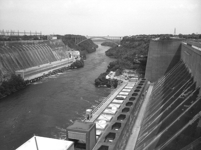 A photograph of two hydroelectric generating stations on either side of the Niagara River. The photo is taken from the south end of Moses Station on the American side of the river, and Beck Stations 1 and 2 can be seen across the river on the Canadian side. A cantilever bridge spans the river in the background.