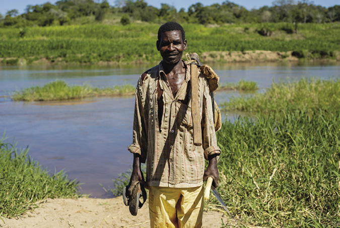 Colour photograph: Photo of a farmer standing in front of a river, cutlass in hand and tools slung over his shoulder.