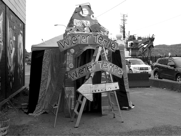 Black and white photograph, a tall square tent sits in a carpark beside a building, a stepladder with elaborate signage repeating “Weiner Temple” twice and an arrow stands in front of the tent, cars and a large ship are in the background.