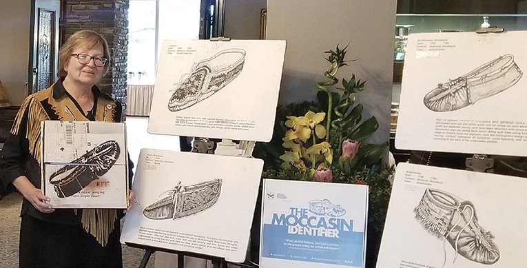 Colour photograph. A woman is holding an image of a moccasin, standing next to four large posters showing detailed drawings of different moccasins. She has short red-brown hair and is wearing glasses and a fringed buckskin vest over a black outfit. In the centre, a label reads “The Moccasin Identifier.” A large flower arrangement is behind this.