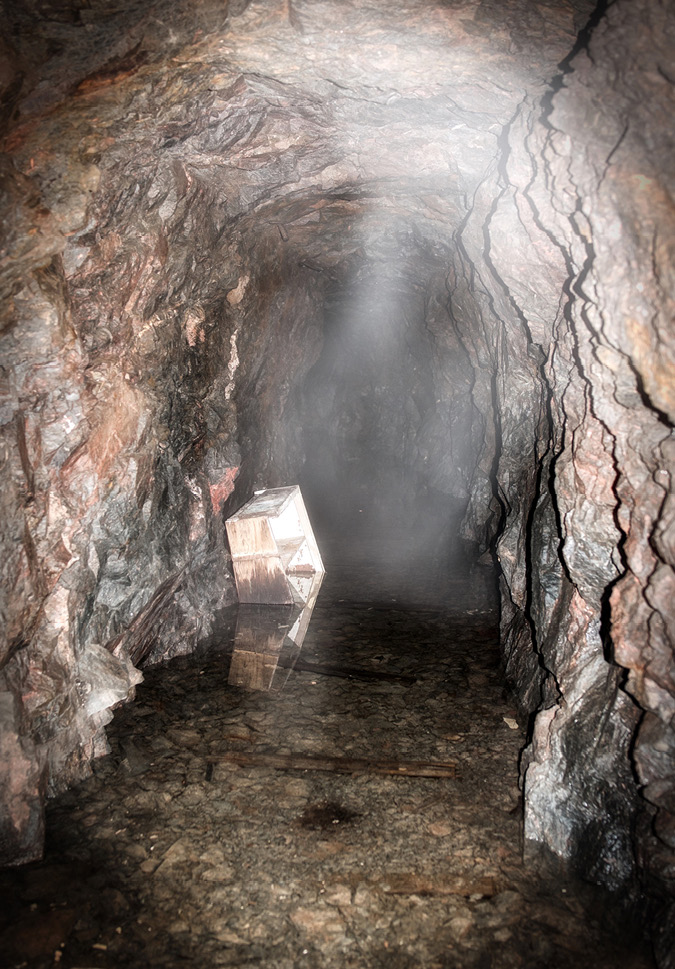 The rocky, damp opening of an old mine shaft has an old structure leaning against the rock wall. Light streaming in shows the glistening damp and mist.