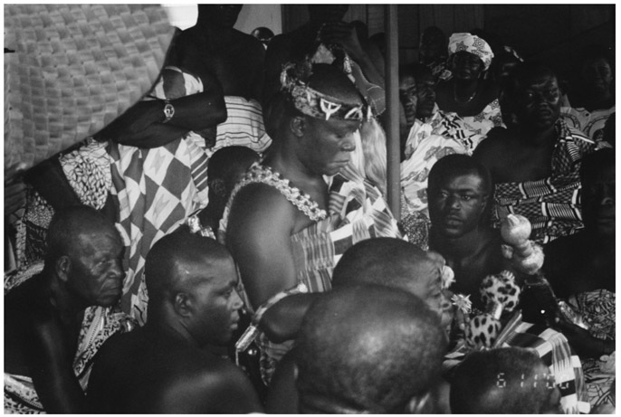 A black and white photograph of Asante King Osei Tutu Two, surrounded by people as he sits at his palace in Kumasi, Ghana. All are dressed in traditional African attire.