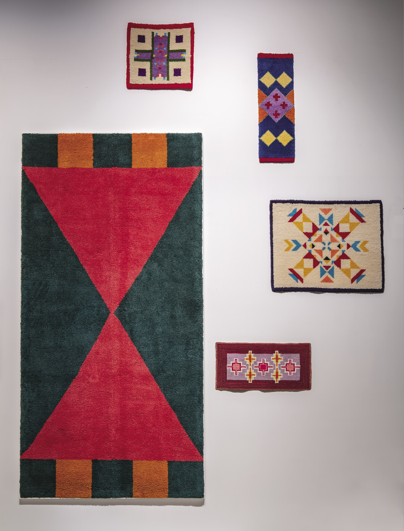 Photo: a white wall with one large red-green hanging surrounded by four much smaller hangings with complex patterns. 