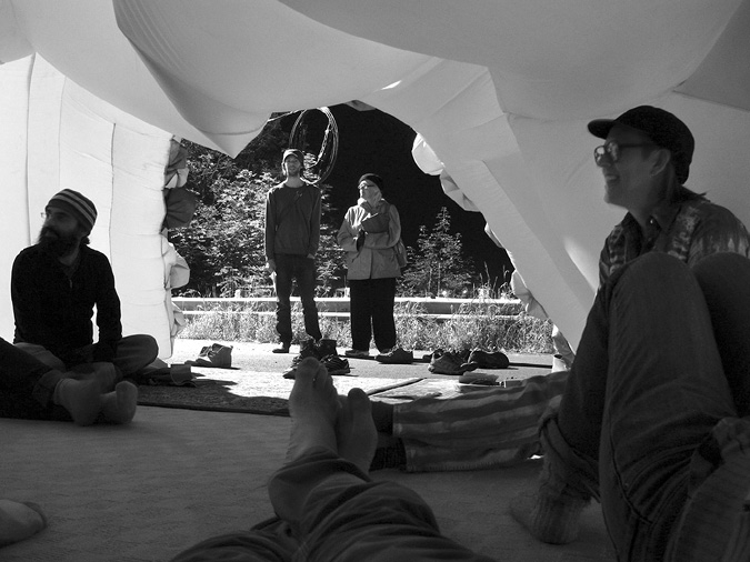 Black and white photograph, a first-person view looks out from inside a white tent door, the photographer’s legs and feet stretch out from the centre of the image, two people stand outside the tent door, while two people and several more legs and feet are seen sitting in the tent; a wide variety of shoes sit at the tent door.