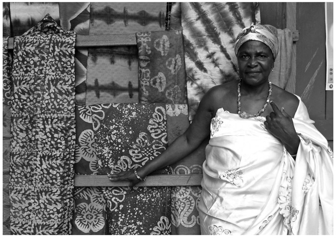 A black and white photograph of Manye Nartekie, Deputy Paramount Queenmother, standing in front of cloth prints on display.