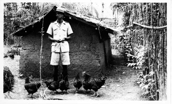 A black and white photograph shows Felix Dibia standing in front of a hut and feeding the chickens. He is wearing a uniform shirt, trouser, and hat.
