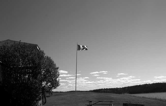 Black and white photograph: A flagpole stands on a grassy hill displaying ACFN’s flag which is blowing in the wind. The flag design has dark panels on the left and the right and is a larger white panel in the centre, displaying a circular logo in the middle. To the left of the flagpole is a large bush concealing a building. To the right is the boreal tree line. The sky is open and clear, with small clouds on the horizon. A body of water flows in the bottom right corner. In the foreground is a picnic table.