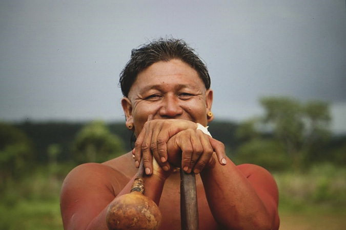 Cacique Jurandir Siridiwe Xavante, leader of the Etenhiritipá Village is standing, holding the top of large stick with one hand on top of the other. His hands are in front of his face, his eyes reveal that he is smiling broadly. The leader is close up and clear in the photo, with his village in the distance in a hazy camera blur.