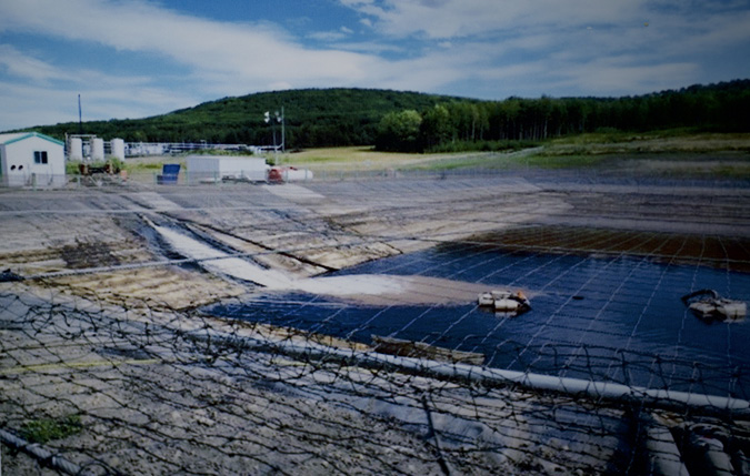 Produced water is stored in a cement dugout in the foreground. To the left side of the dugout are white buildings, electric set up and storage. Large metal pumping equipment sit in the middle of the water storage dugout, moving the water.  The dugout has a netting cover that is rested on top of the dugout.