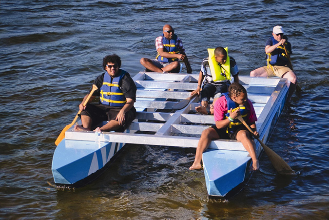 Five people wearing lifejackets are seated on the top canopy of the Art Ark, a combination of boats and canopy. Two of the people on the boar project are paddling in a body of water with oars.