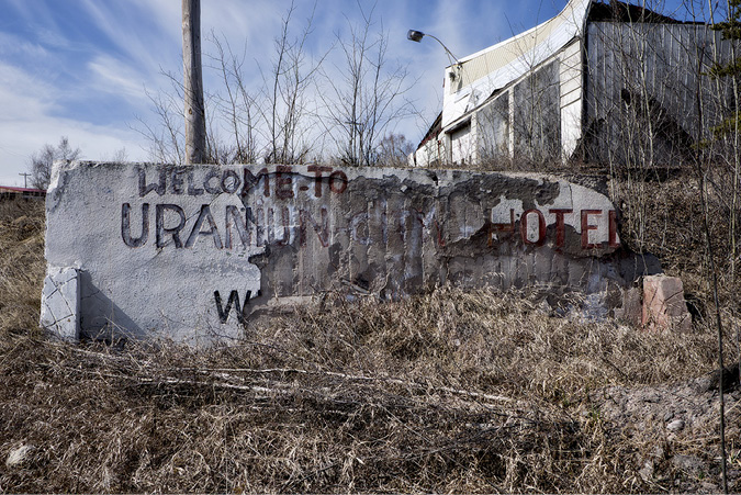 An old welcome sign to the Uranium City hotel is now broken, chipped and sitting in the grass, in the background is an old dilapidated building.